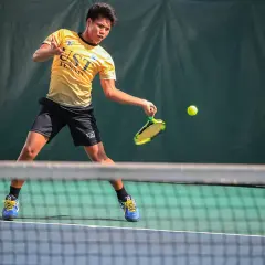 Młody tenisista w żółtej koszulce UST TENNIS uderza piłkę, walcząc o każdy break point.