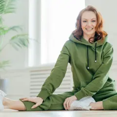 Uśmiechnięta rudowłosa kobieta w zielonym dresie, który wygląda na wykonany z miękkiego **cottonu**. Rozciąga się na podłodze obok rośliny.