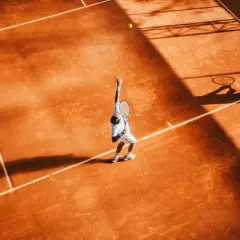 Agassi tenisista w akcji, serwuje na ceglanym korcie. Cień rakiety i sylwetka gracza rzucają się na pomarańczową nawierzchnię.