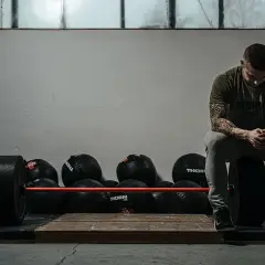 Mężczyzna z tatuażami siedzi na talerzu obciążeniowym, gotowy do treningu crossfit w domu. Obok sztanga i piłki lekarskie.