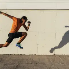 Biegacz w pomarańczowej koszulce i czarnych spodenkach w dynamicznym skoku, jego cień odbija się na ścianie. Trening na świeżym powietrzu, by poprawić VO2 max.