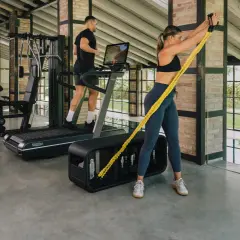 Kobieta wykonuje trening funkcjonalny z gumą oporową, mężczyzna biegnie na bieżni w nowoczesnej siłowni.