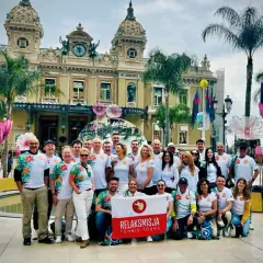 Grupa turystów z flagą "RelaksMisja Tennis Tours" przed kasynem w Monte Carlo. Wszyscy ubrani w koszulki z motywem kwiatowym.