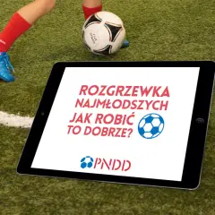 Tablet na boisku z napisem "Rozgrzewka najmłodszych jak robić to dobrze?". Obok piłka i noga dziecka w czerwonej skarpecie.
