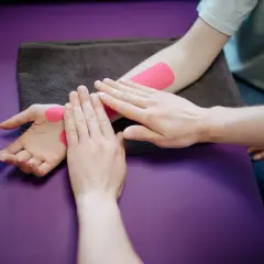 Fizjoterapeuta wykonuje slump test, aplikując różową taśmę kinezjotaping na przedramieniu i dłoni pacjenta.