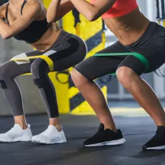 Dwie kobiety wykonują przysiady z gumami oporowymi na udach. Ćwiczenie to angażuje mięśnie podobnie jak maszyna hack squat, wzmacniając nogi i pośladki.