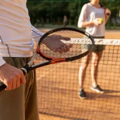 Mężczyzna trzyma rakietę tenisową, czując jej siłę naciągu. W tle kobieta z piłką czeka na grę.