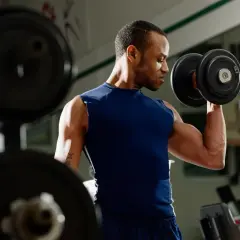 Mężczyzna ćwiczy biceps z hantlem. W tle widać sprzęt do ćwiczeń.