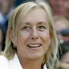 Uśmiechnięta Martina Navratilova w białej marynarce, z blond włosami i kolczykami w kształcie kółek, obserwuje wydarzenie.