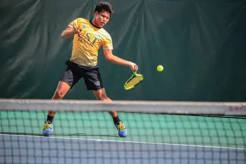 Młody tenisista w żółtej koszulce UST TENNIS uderza piłkę, walcząc o każdy break point.