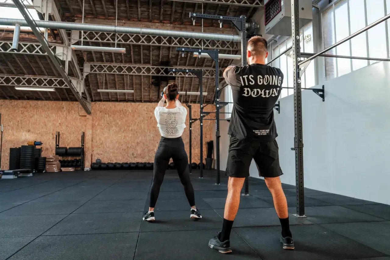 Para pozuje na siłowni. Ona wykonuje ćwiczenie z kettlebell, on stoi z tyłu. To właśnie crossfit co to za trening!