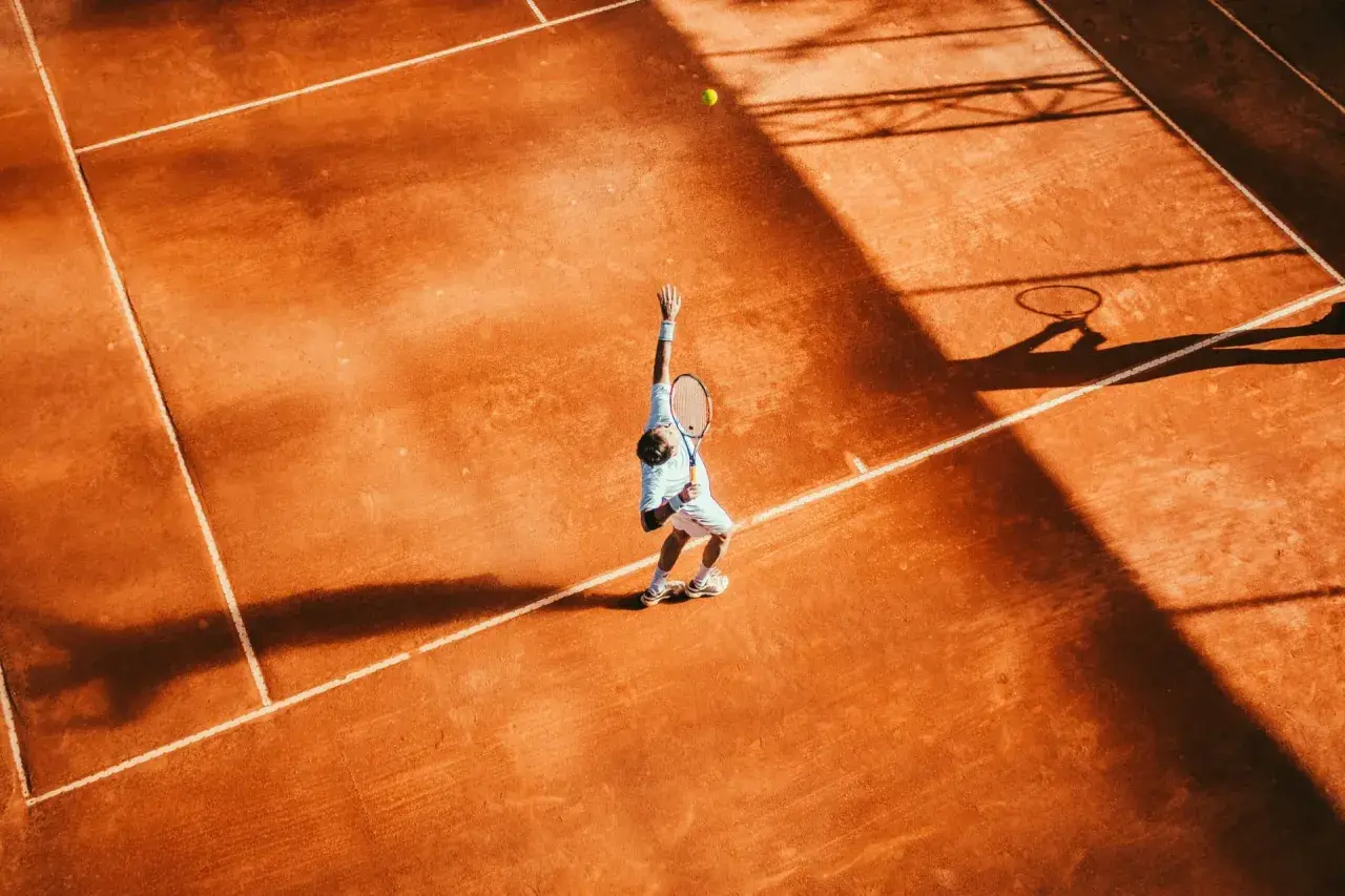 Agassi tenisista w akcji, serwuje na ceglanym korcie. Cień rakiety i sylwetka gracza rzucają się na pomarańczową nawierzchnię.