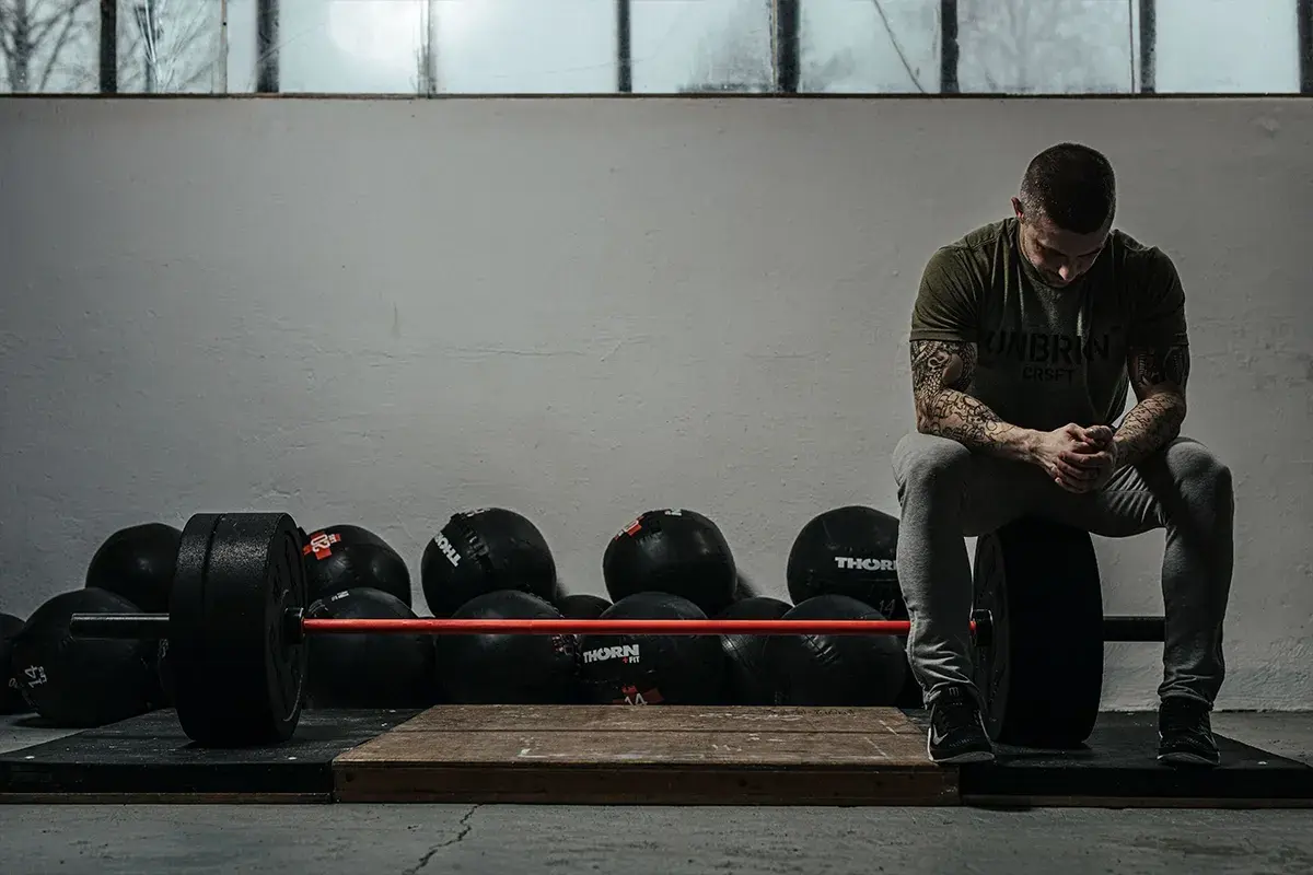 Mężczyzna z tatuażami siedzi na talerzu obciążeniowym, gotowy do treningu crossfit w domu. Obok sztanga i piłki lekarskie.