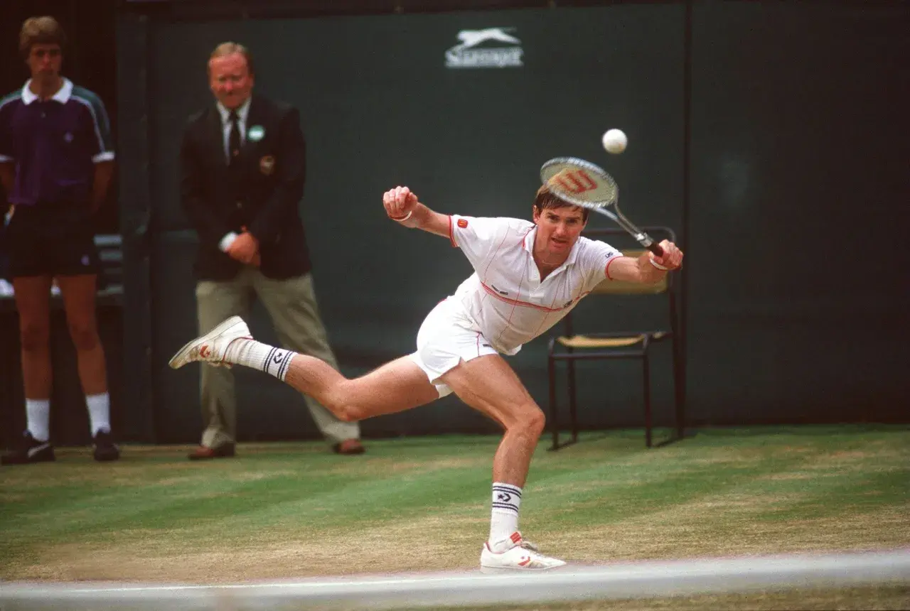 Jimmy Connors w akcji, uderza piłkę na trawiastym korcie. W tle sędzia i chłopiec podający piłki.
