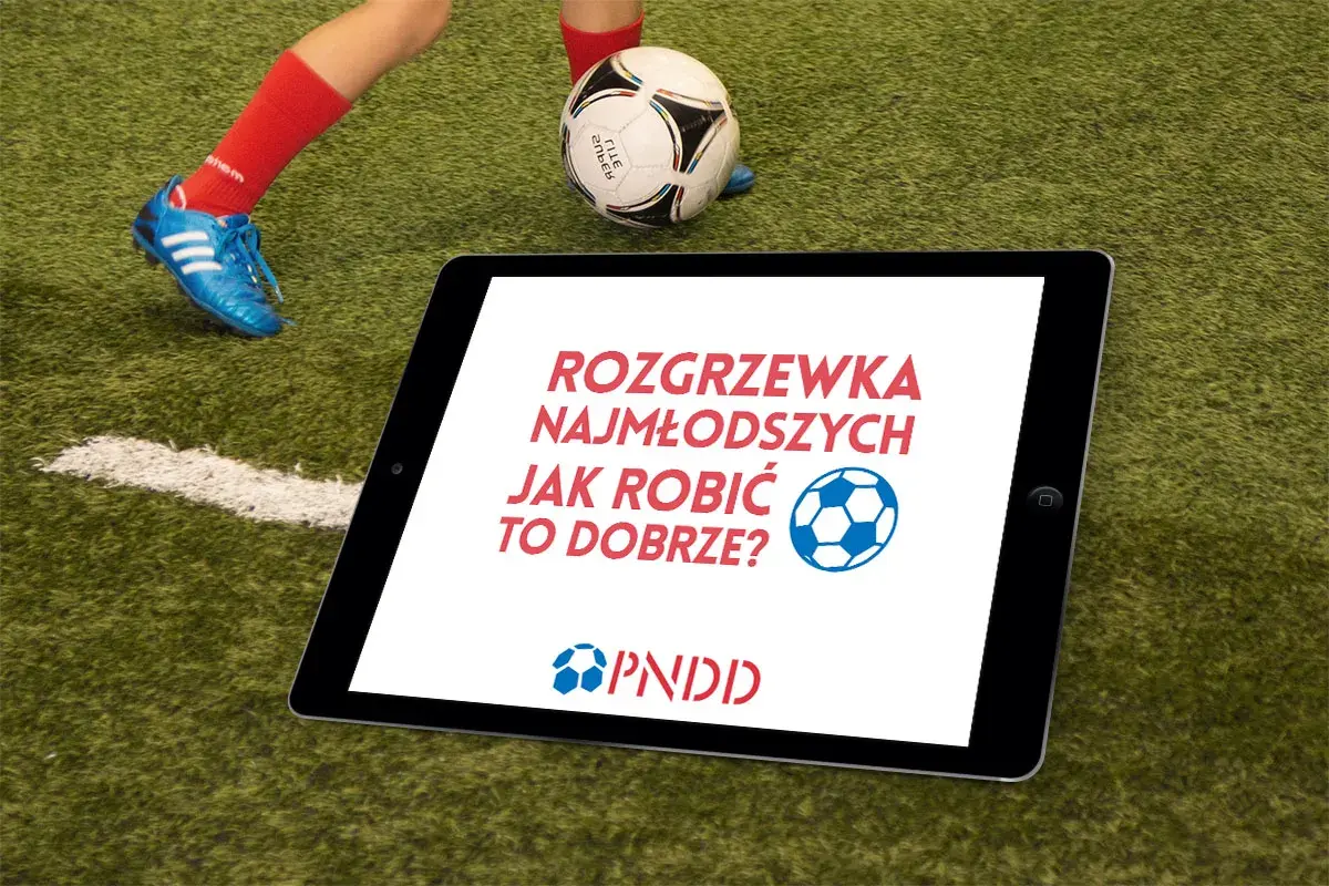 Tablet na boisku z napisem "Rozgrzewka najmłodszych jak robić to dobrze?". Obok piłka i noga dziecka w czerwonej skarpecie.