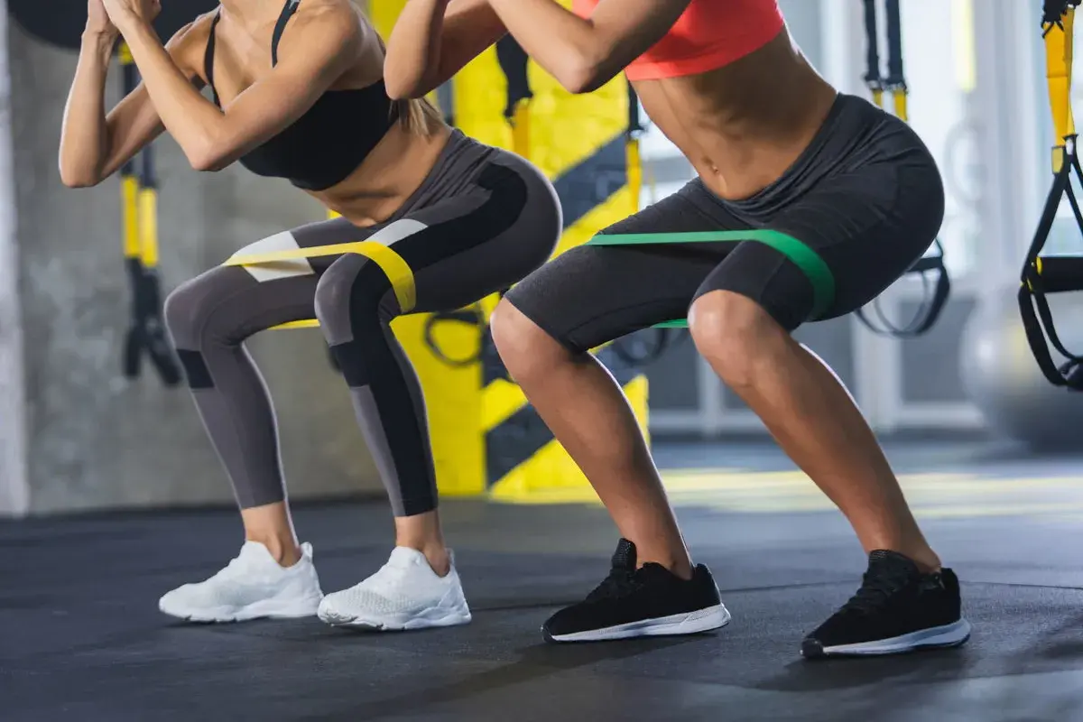 Dwie kobiety wykonują przysiady z gumami oporowymi na udach. Ćwiczenie to angażuje mięśnie podobnie jak maszyna hack squat, wzmacniając nogi i pośladki.