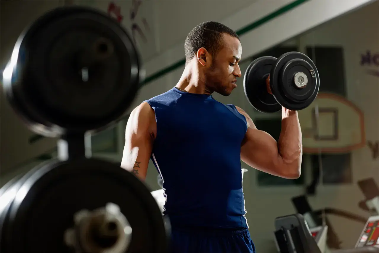Mężczyzna ćwiczy biceps z hantlem. W tle widać sprzęt do ćwiczeń.