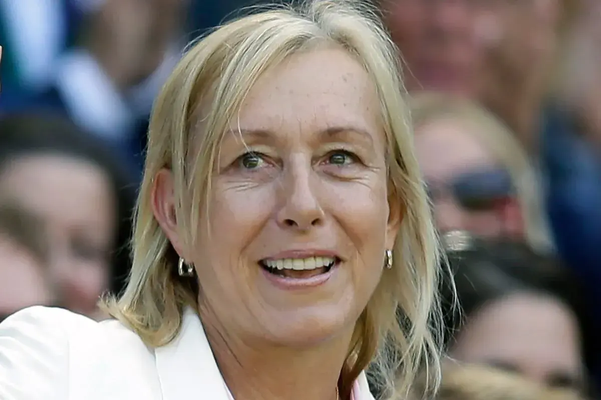 Uśmiechnięta Martina Navratilova w białej marynarce, z blond włosami i kolczykami w kształcie kółek, obserwuje wydarzenie.