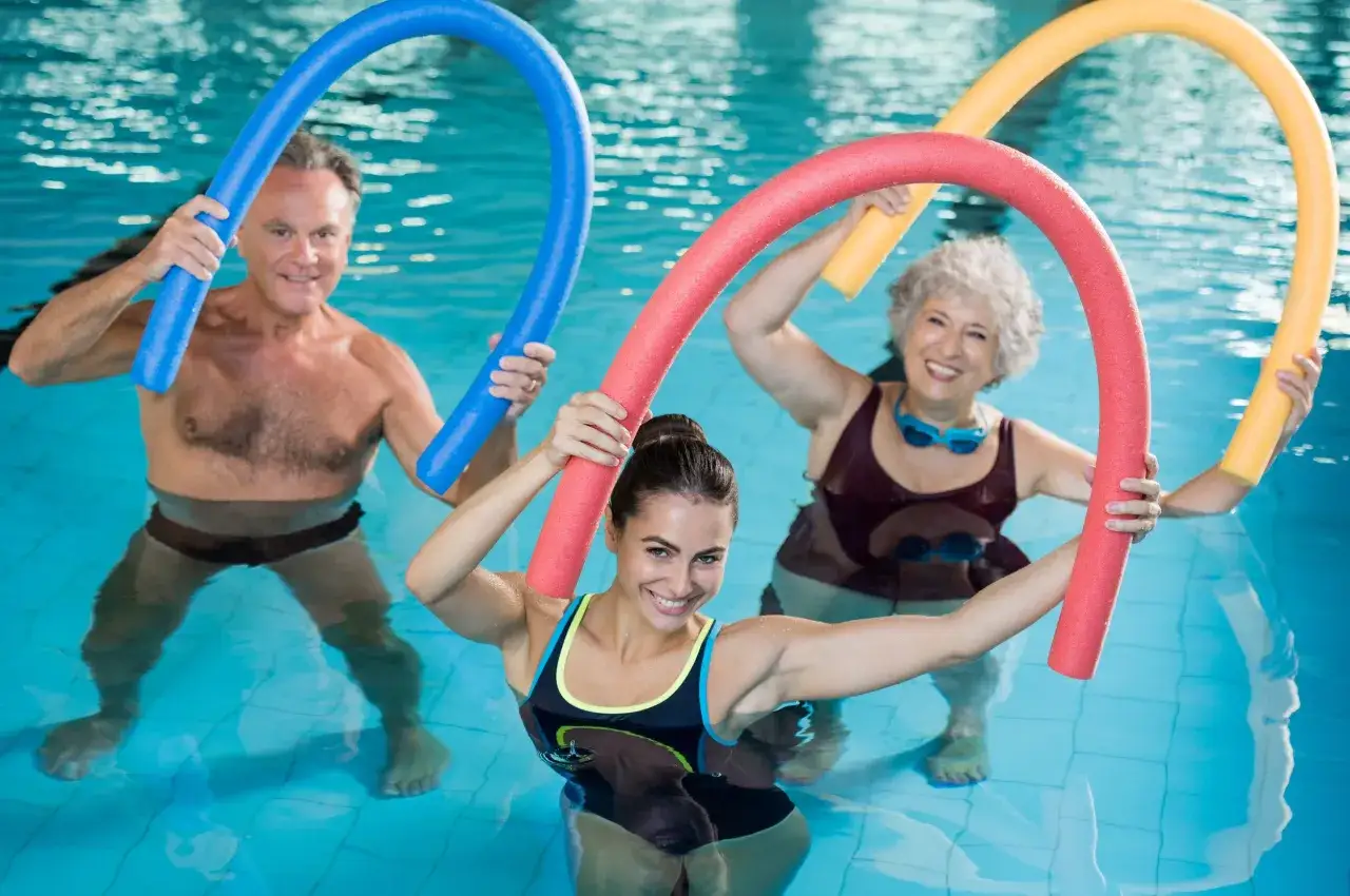 Uśmiechnięci ludzie ćwiczą aqua fitness z kolorowymi makaronami pływackimi w basenie.