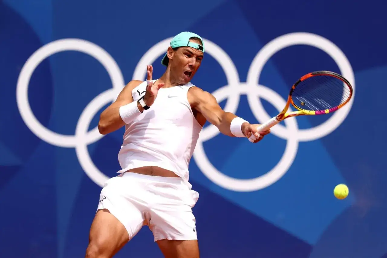Rafael Nadal w akcji na korcie, w tle olimpijskie kółka. Ile lat ma Rafael Nadal? To pytanie nurtuje fanów tenisa.