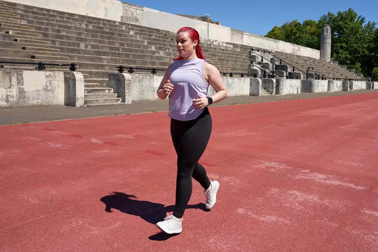 Kobieta z czerwonymi włosami biegnie po czerwonym torze lekkoatletycznym. To jej test coopera, przygotowuje się do zawodów.