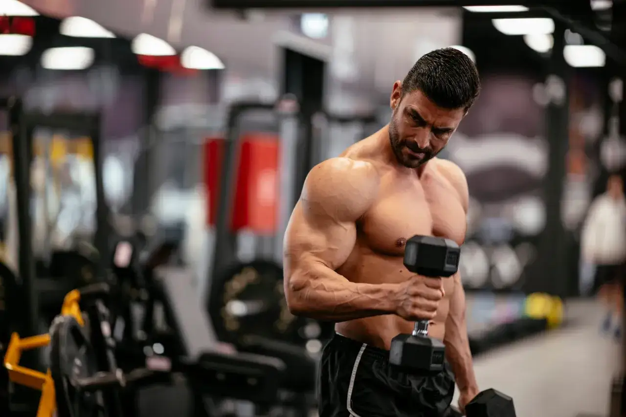 Mężczyzna z imponującymi mięśniami ćwiczy pompki na biceps z hantlami w siłowni.
