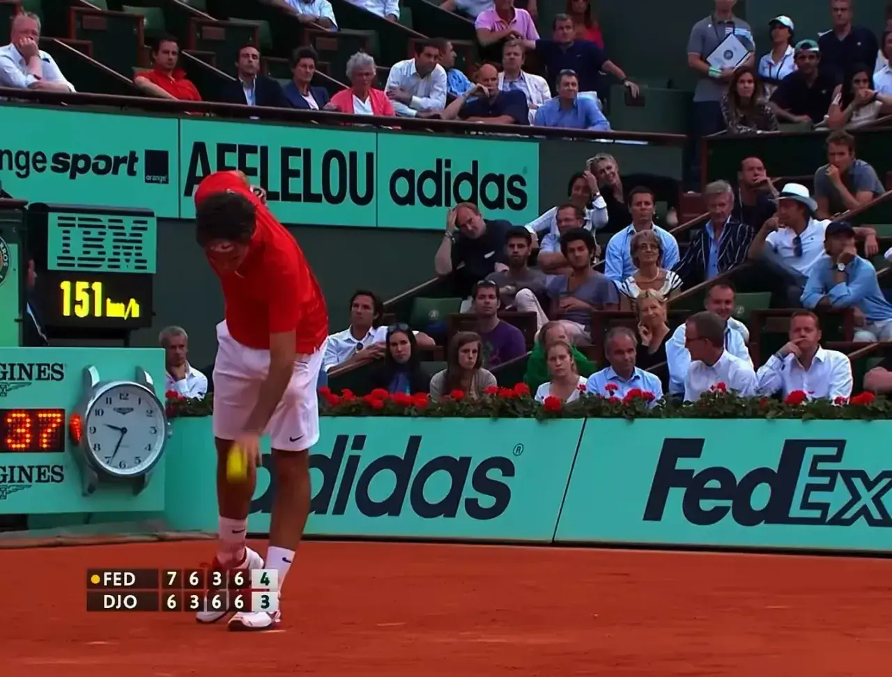 Tenisista Federer w czerwonej koszulce serwuje piłkę na korcie ziemnym. Widownia ogląda mecz.
