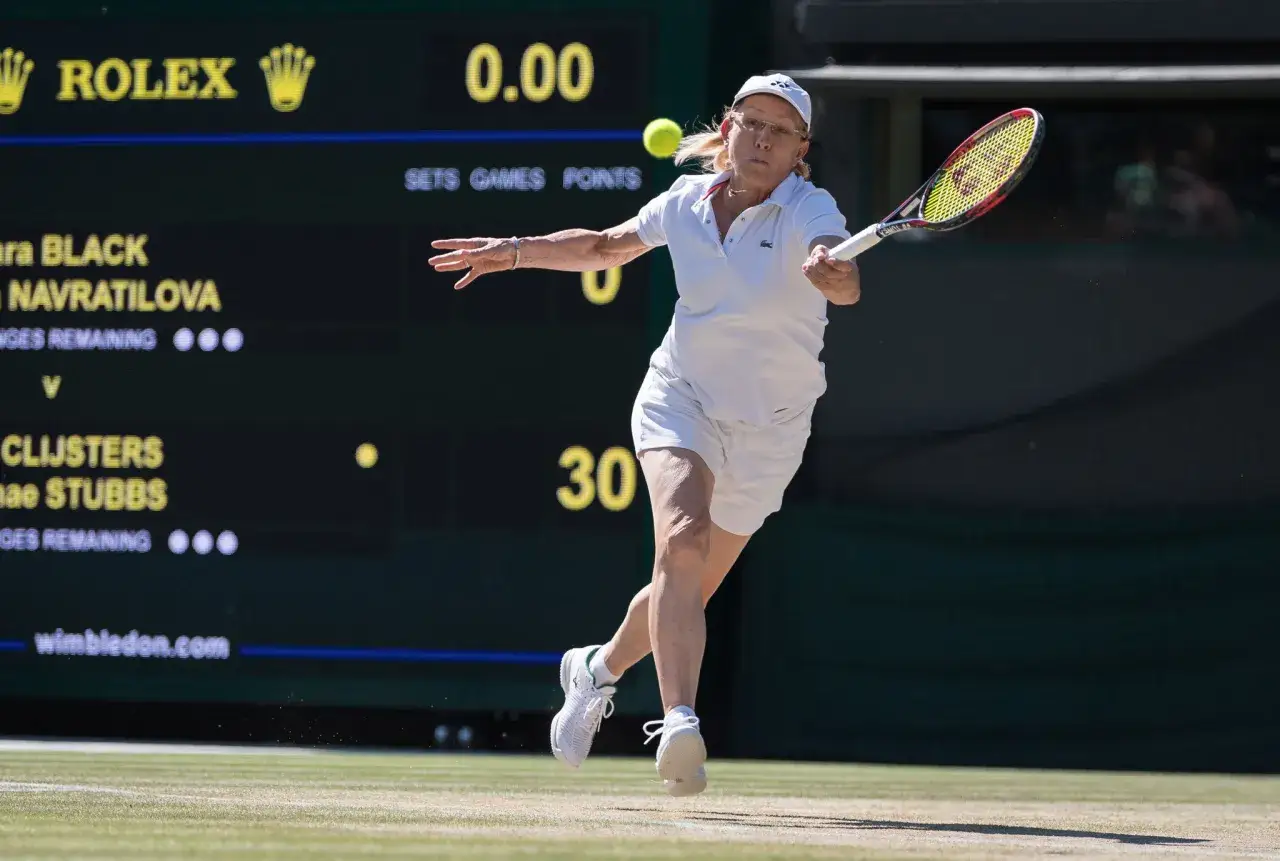 Martina Navratilova w akcji na korcie, w białym stroju, z rakietą w dłoni. Na tablicy wyników widać jej nazwisko.